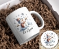 Preview: Weihnachtstasse mit fröhlichem Schneemann-Motiv und passendem Untersetzer: Ein lächelnder Schneemann mit roter Mütze und Schal hebt fröhlich die Arme, umgeben von Schneeflocken und winterlichen Details. Unter dem Motiv steht der Schriftzug ‚Let it Snow‘.
